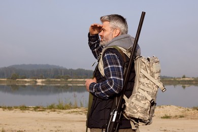 Photo of Man with hunting rifle and backpack near lake outdoors. Space for text