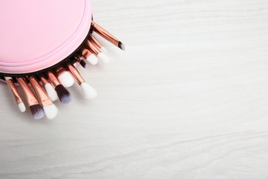 Photo of Bag with set of professional makeup brushes on light wooden table, above view. Space for text
