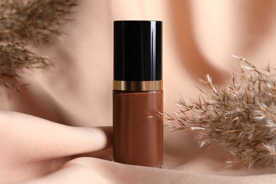 Photo of Bottle of skin foundation and dried reeds on beige cloth, closeup. Makeup product