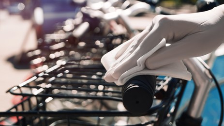 Photo of Woman in latex gloves cleaning bicycle handlebar with wet wipe outdoors, closeup. Protective measures