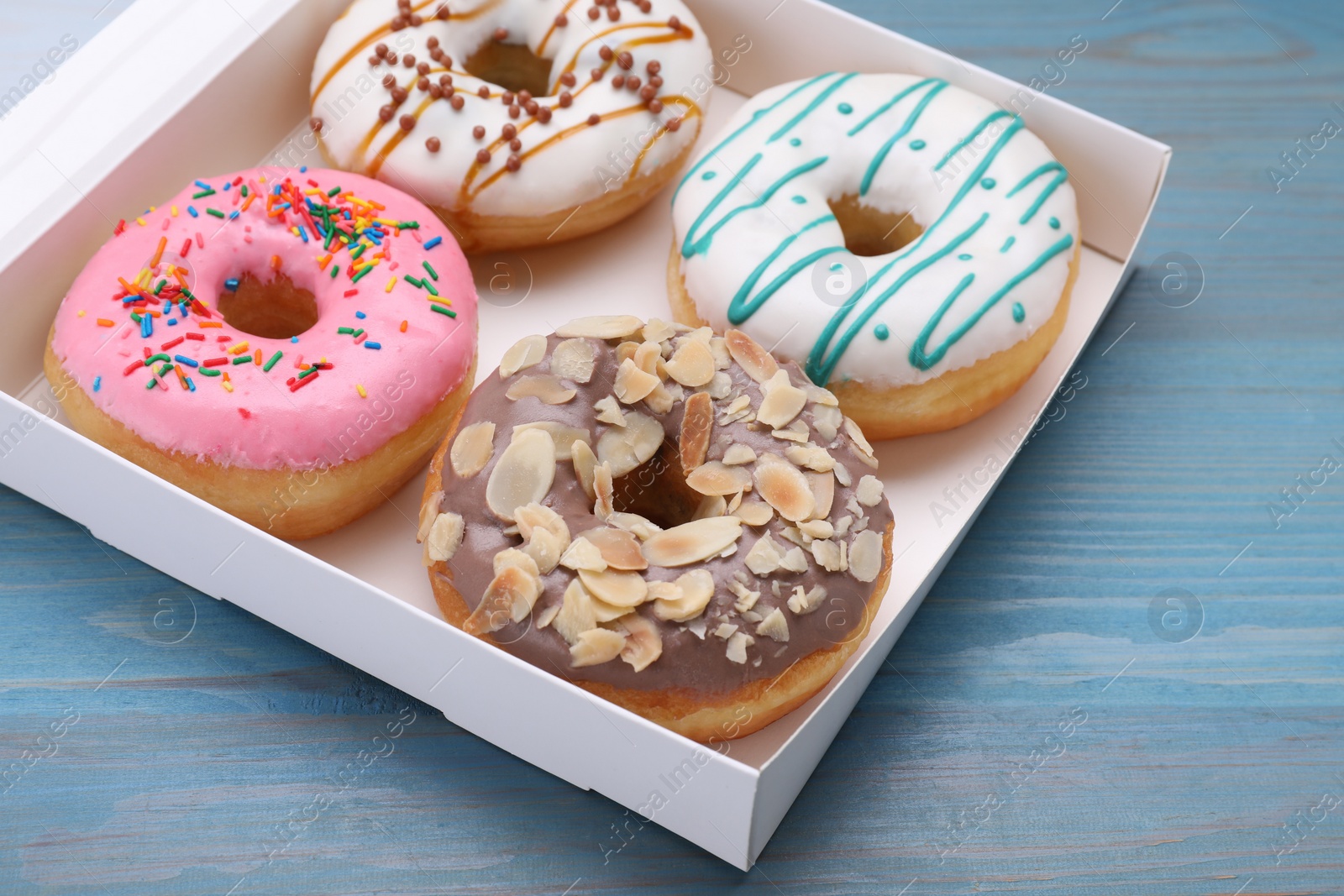 Photo of Box with different tasty glazed donuts on light blue wooden table, closeup