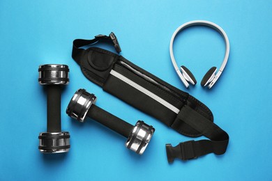 Photo of Stylish black waist bag, dumbbells and headphones on light blue background, flat lay
