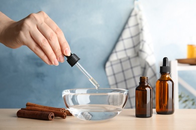 Photo of Woman dripping cinnamon essential oil from pipette into bowl of water on table, closeup