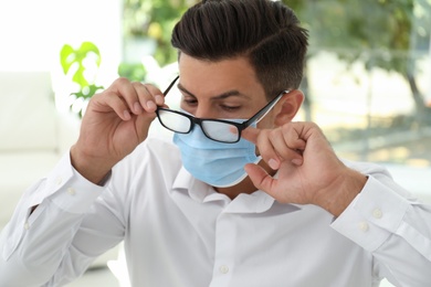 Photo of Man wiping foggy glasses caused by wearing medical mask indoors