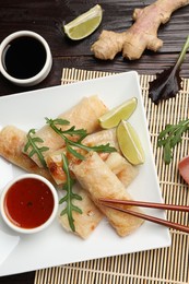 Photo of Tasty fried spring rolls, arugula, lime and sauces served on wooden table, flat lay