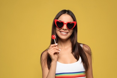 Portrait of beautiful young woman with heart shaped sunglasses and lollipop on color background