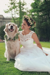 Photo of Bride and adorable Golden Retriever wearing wreath made of beautiful flowers on green grass outdoors