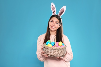 Photo of Beautiful woman in bunny ears headband holding basket with Easter eggs on color background