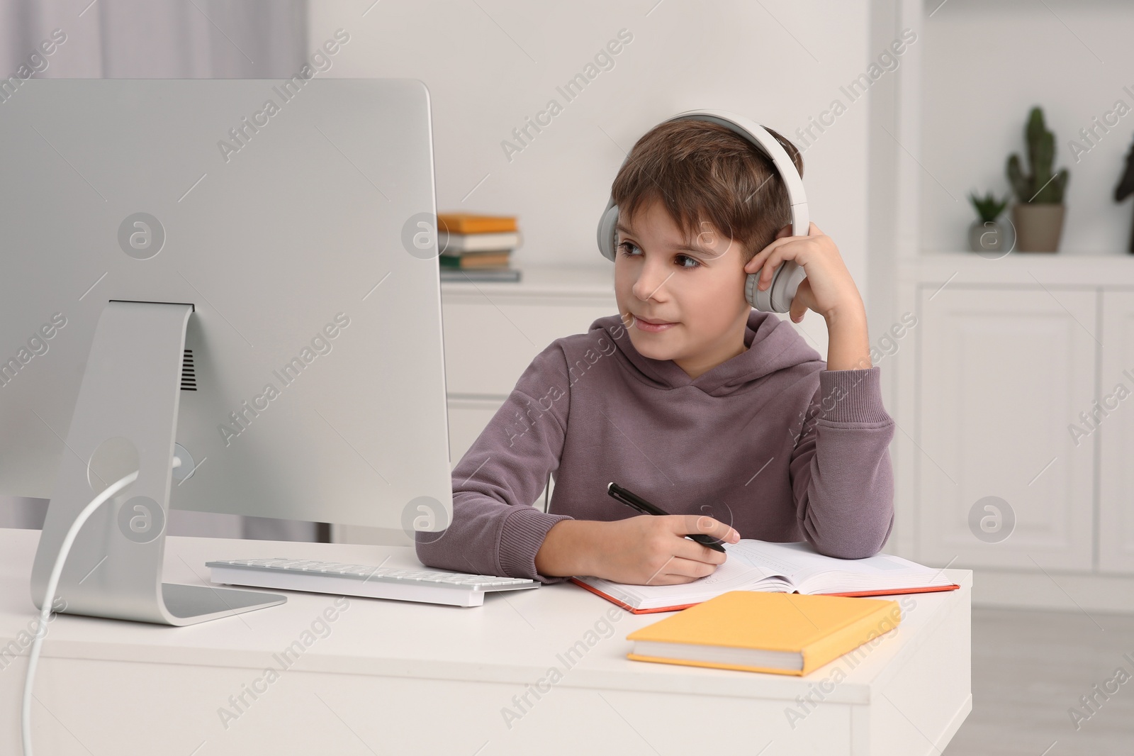 Photo of Boy writing in notepad while using computer and headphones at desk in room. Home workplace