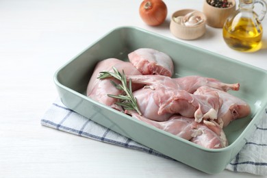 Photo of Raw rabbit meat and rosemary in baking dish on white wooden table, closeup