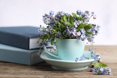 Photo of Beautiful forget-me-not flowers in cup, saucer and books on wooden table against light background, closeup