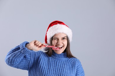 Photo of Pretty woman in Santa hat and blue sweater eating candy cane on grey background