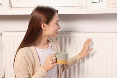 Photo of Woman holding cup with hot drink and warming hand on heating radiator indoors