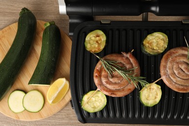 Photo of Electric grill with homemade sausages, zucchini and rosemary on wooden table, flat lay