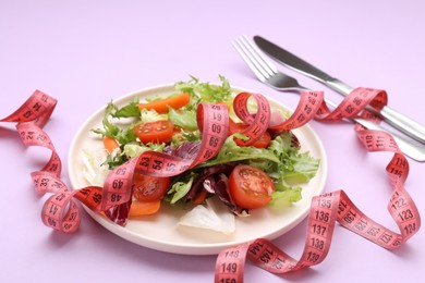 Photo of Plate with fresh vegetable salad and measuring tape on violet background, closeup. Healthy diet concept