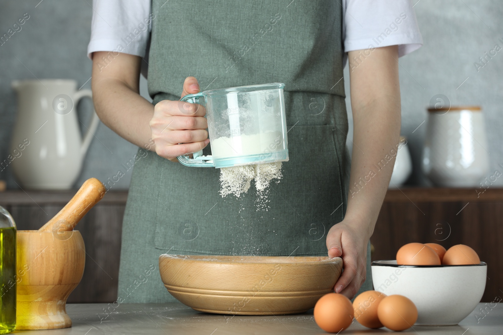 Photo of Woman sieving flour into bowl at table in kitchen, closeup