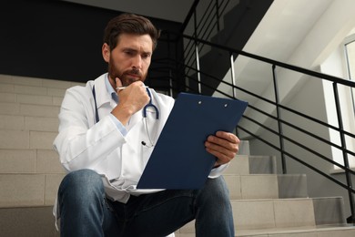 Photo of Pensive doctor with clipboard sitting on stairs in hospital