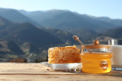 Photo of Fresh aromatic honey, combs and glass of milk on wooden table against mountain landscape. Space for text