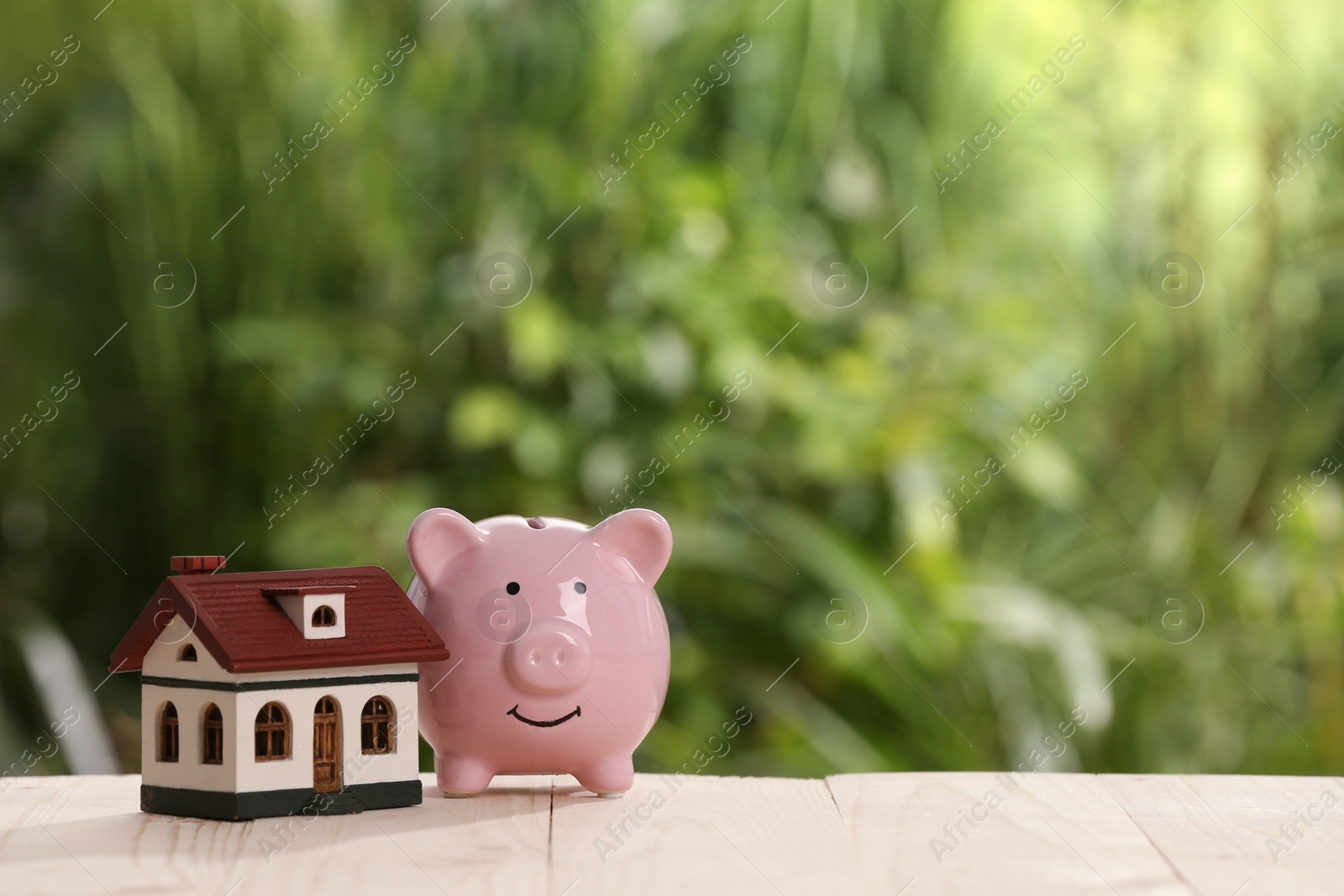 Photo of Piggy bank and little house toy on white wooden table outdoors. Space for text