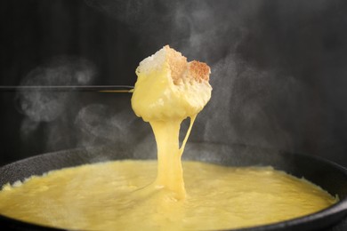 Image of Dipping piece of bread into fondue pot with tasty melted cheese against dark background, closeup