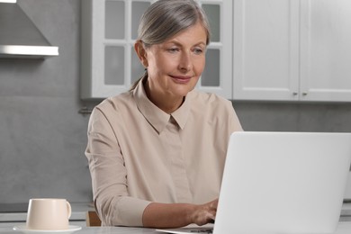 Photo of Beautiful senior woman using laptop at table in kitchen