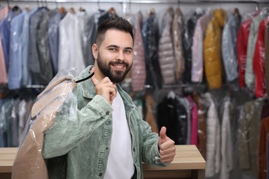Photo of Dry-cleaning service. Happy man holding hanger with coat in plastic bag and showing thumb up indoors, space for text