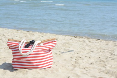 Photo of Stylish striped bag with sunglasses on sandy beach near sea, space for text