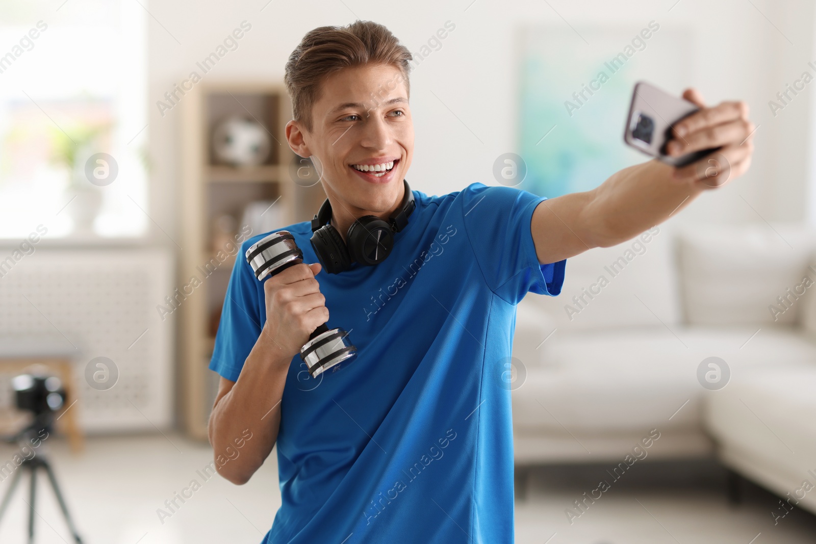 Photo of Smiling sports blogger with dumbbell streaming online fitness lesson with smartphone at home