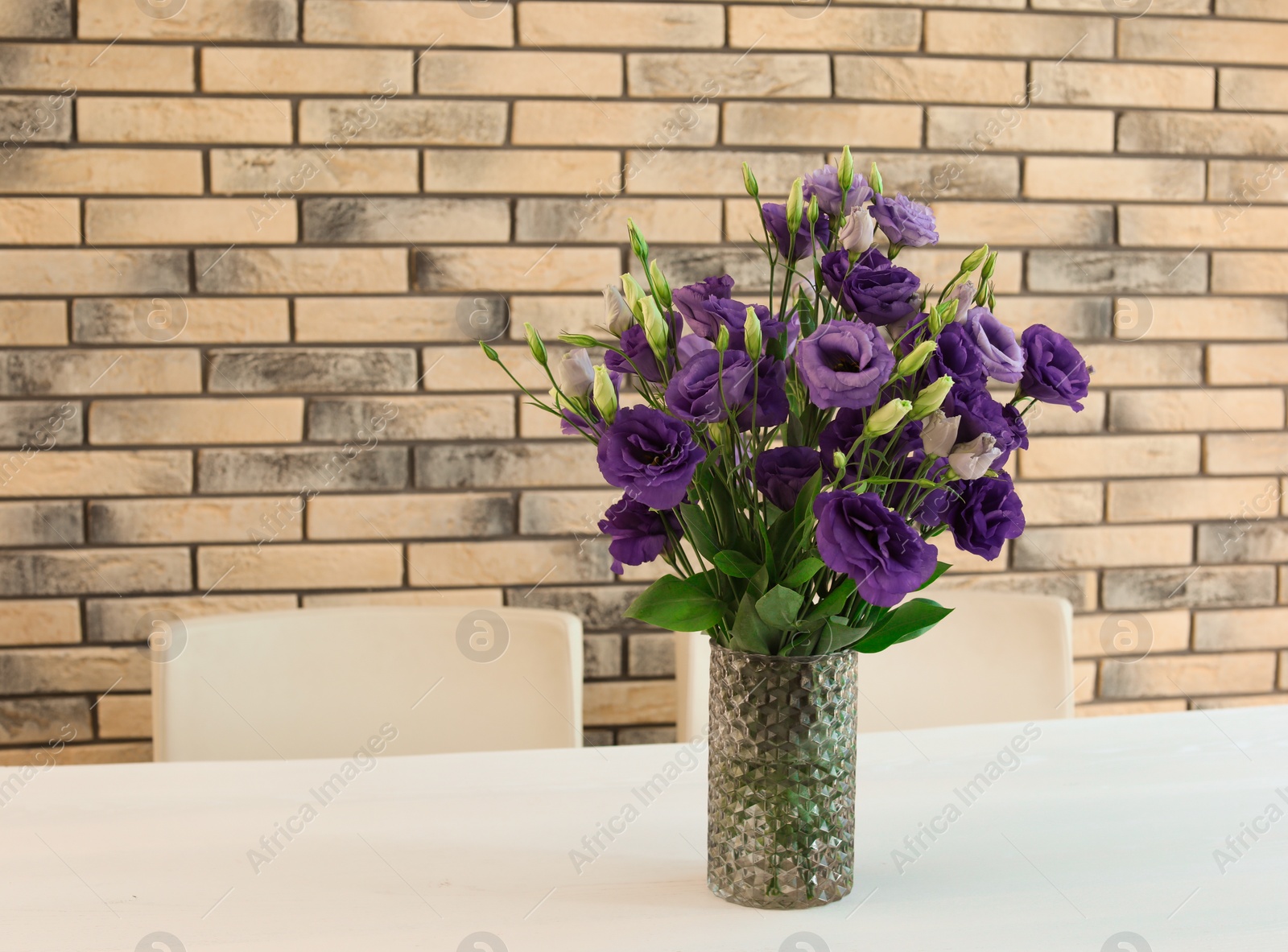 Photo of Vase with beautiful flowers on white table against brick wall. Stylish interior