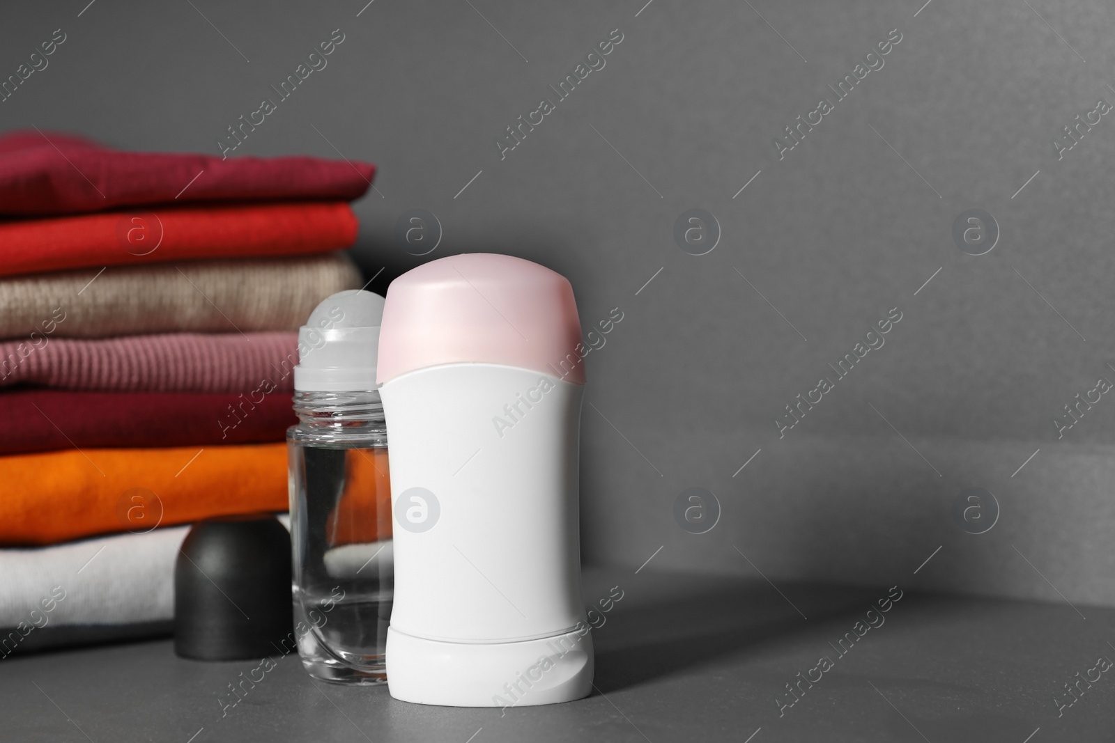 Photo of Deodorants near stack of clean clothes on grey table. Space for text