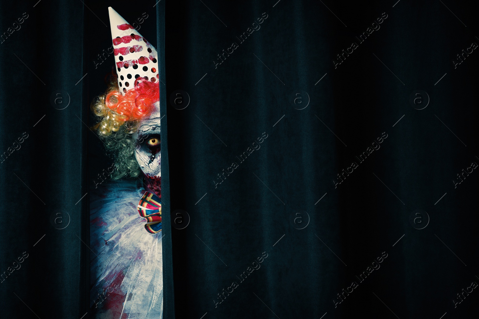 Photo of Terrifying clown hiding behind black curtains, space for text. Halloween party costume