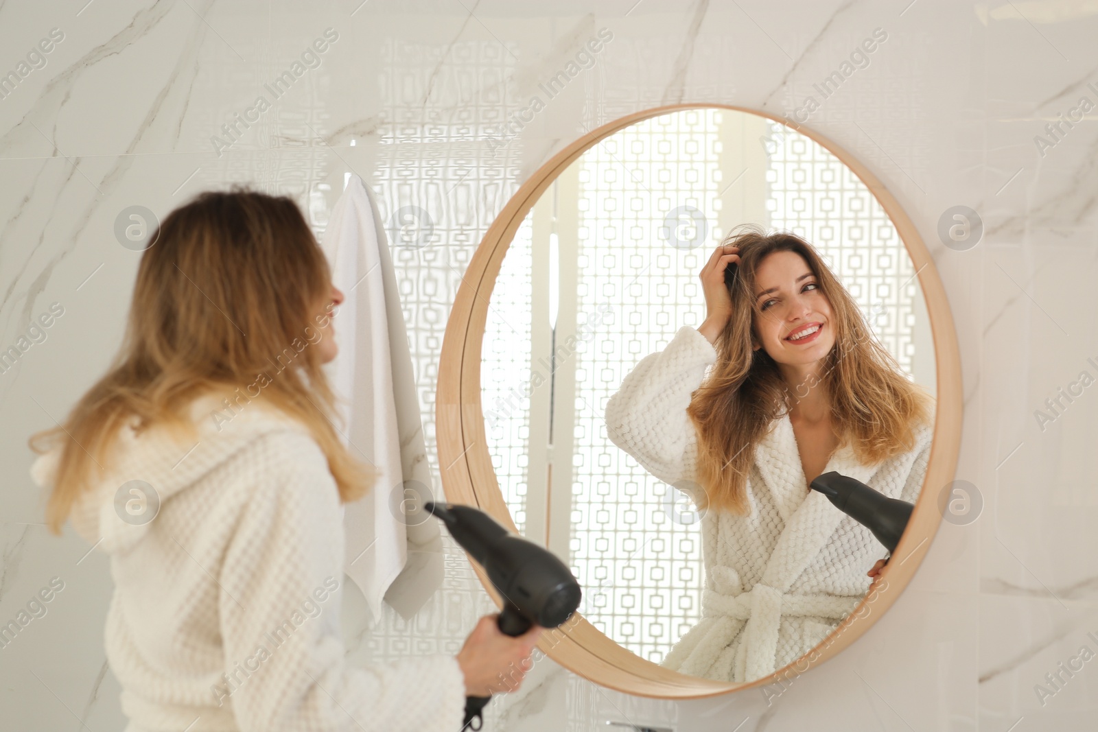 Photo of Beautiful young woman using hair dryer near mirror at home