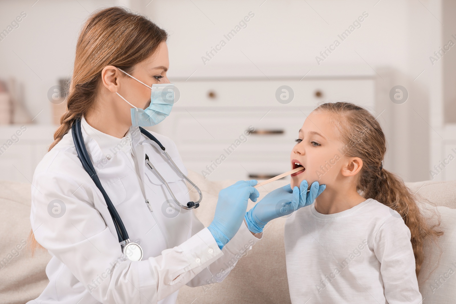 Photo of Doctor in medical mask examining girl`s oral cavity with tongue depressor indoors