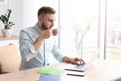 Photo of Young man drinking coffee while working with laptop at desk. Home office