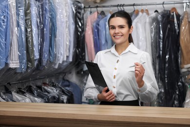 Dry-cleaning service. Happy worker with clipboard and pen at counter indoors, space for text