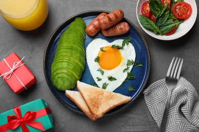 Romantic breakfast with fried heart shaped egg, avocado, sausages and toasts on grey table, flat lay. Valentine's day celebration