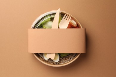 Photo of Tasty food in container with wooden fork and knife on beige background, top view. Space for text