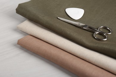Different fabrics, scissors and tailor's chalk on light beige wooden table, closeup