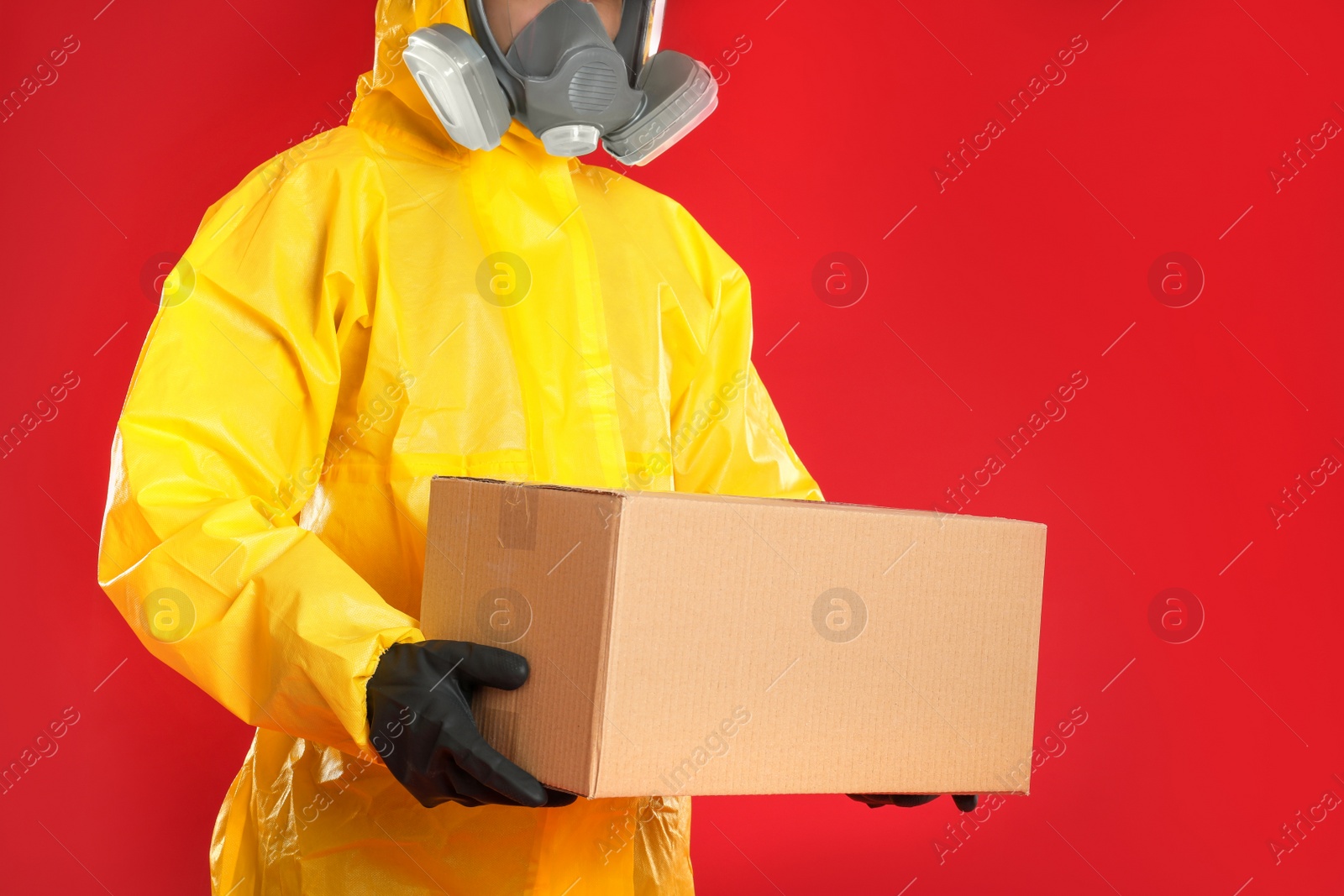 Photo of Man wearing chemical protective suit with cardboard box on red background, closeup. Prevention of virus spread