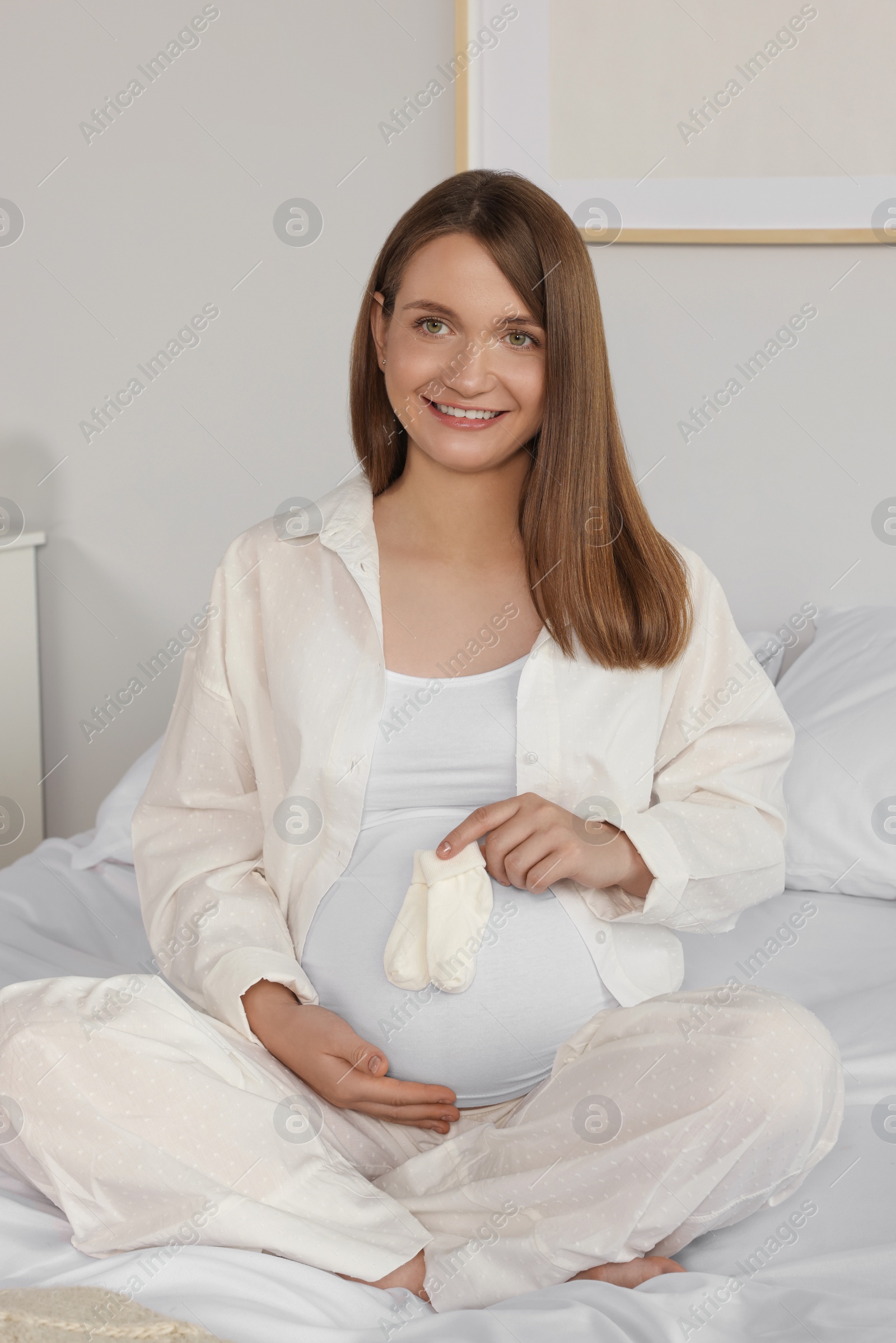 Photo of Happy young pregnant woman with baby socks on bed at home