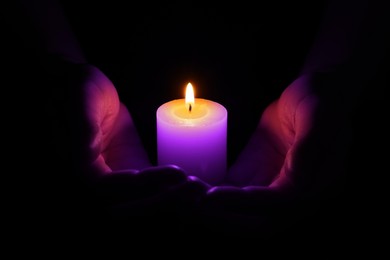 Woman holding burning violet candle in hands on black background, closeup. Funeral attributes