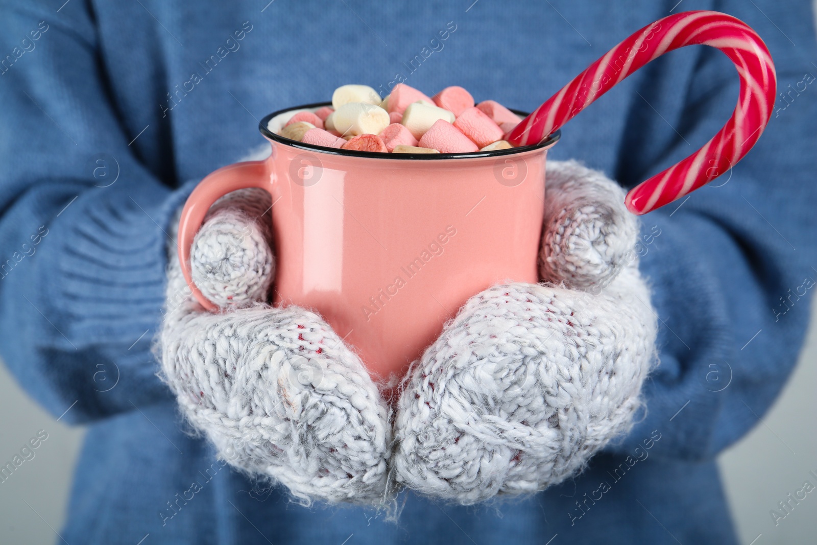 Photo of Woman in knitted mittens holding cup of delicious hot chocolate with marshmallows and candy cane, closeup