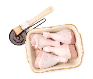 Photo of Raw chicken drumsticks in baking dish, marinade and basting brush isolated on white, top view