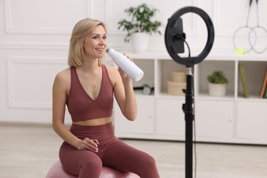 Photo of Smiling sports blogger holding bottle while streaming online fitness lesson at home