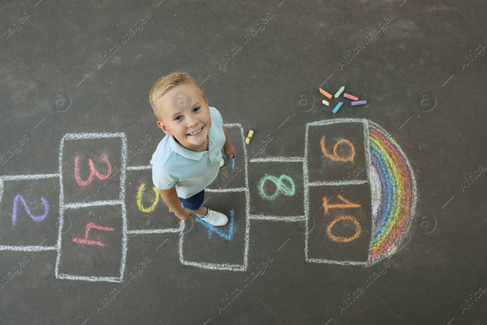 Photo of Little boy and colorful hopscotch drawn with chalk on asphalt outdoors, top view. Happy childhood