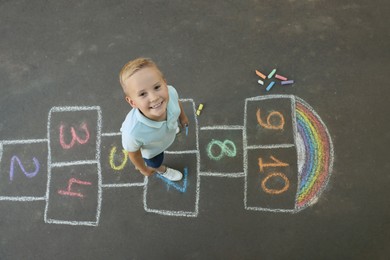 Photo of Little boy and colorful hopscotch drawn with chalk on asphalt outdoors, top view. Happy childhood