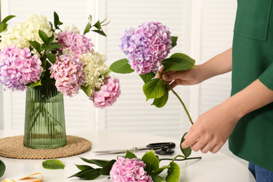 Woman making bouquet with beautiful hydrangea flowers at table indoors, closeup. Interior design element