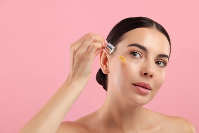 Beautiful young woman applying serum onto her face on pink background