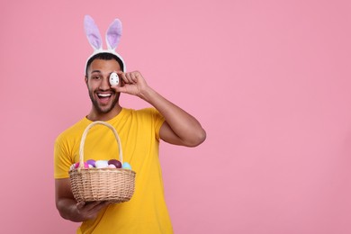 Photo of Happy African American man in bunny ears headband covering eye with Easter egg on pink background, space for text