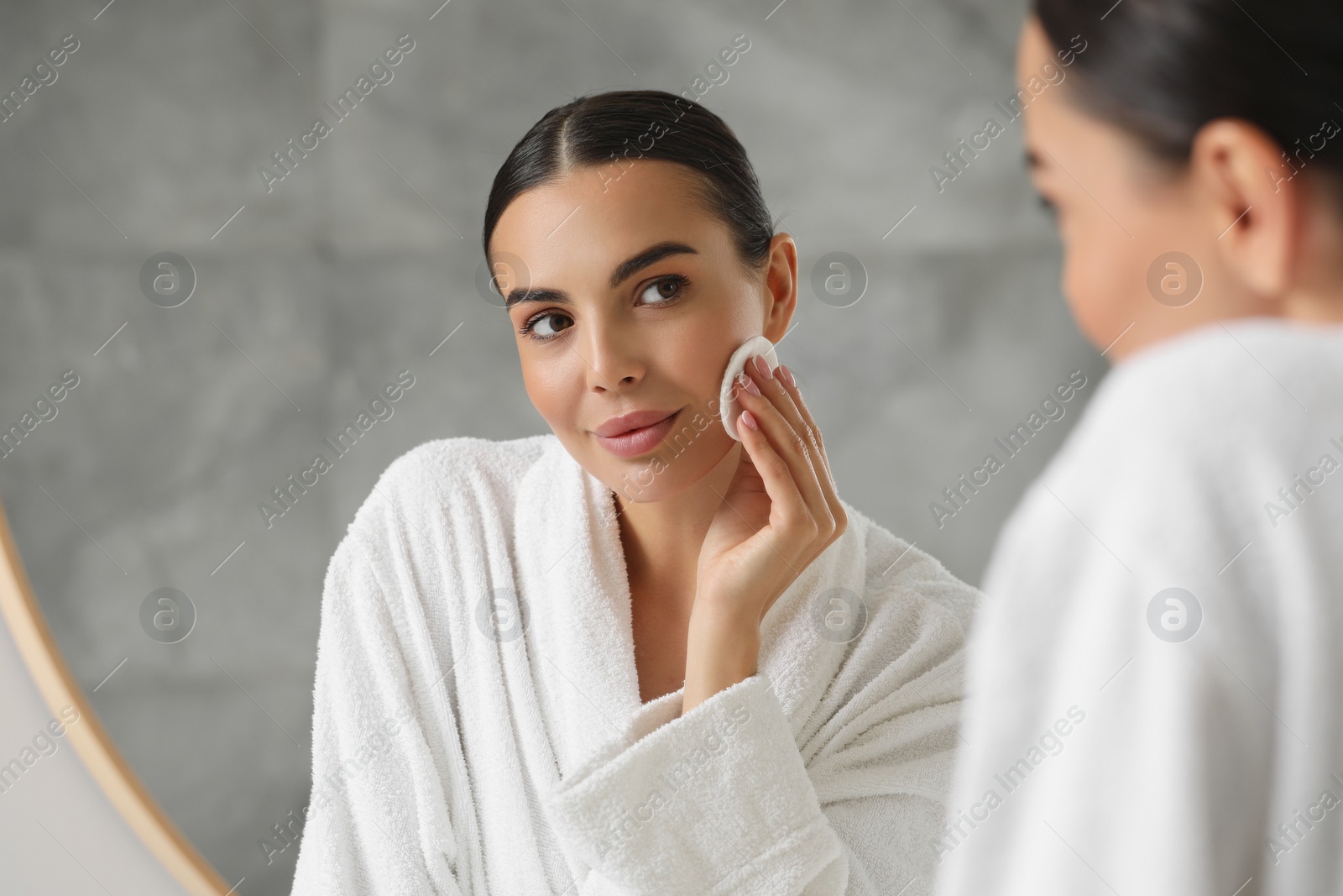 Photo of Beautiful woman removing makeup with cotton pad near mirror indoors